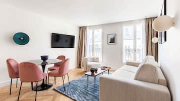 A modern living room with a sofa, a dining table with chairs, a flat-screen TV, and a large window with curtains.