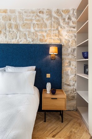 Una camera da letto con parete in pietra, una testiera blu, un comodino in legno con una lampada e un letto bianco.