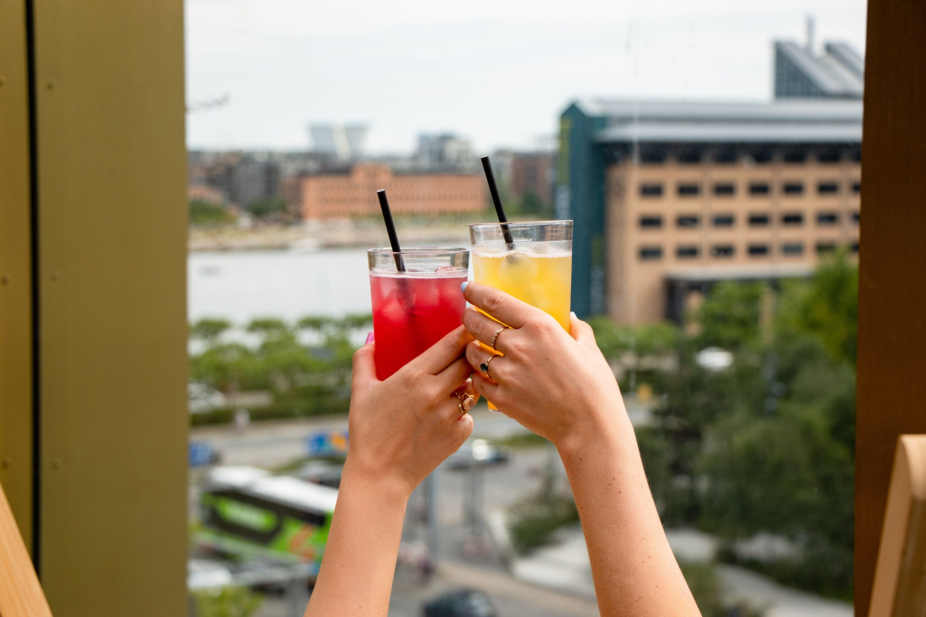 Two hands holding glasses of beverages with a cityscape in the background.