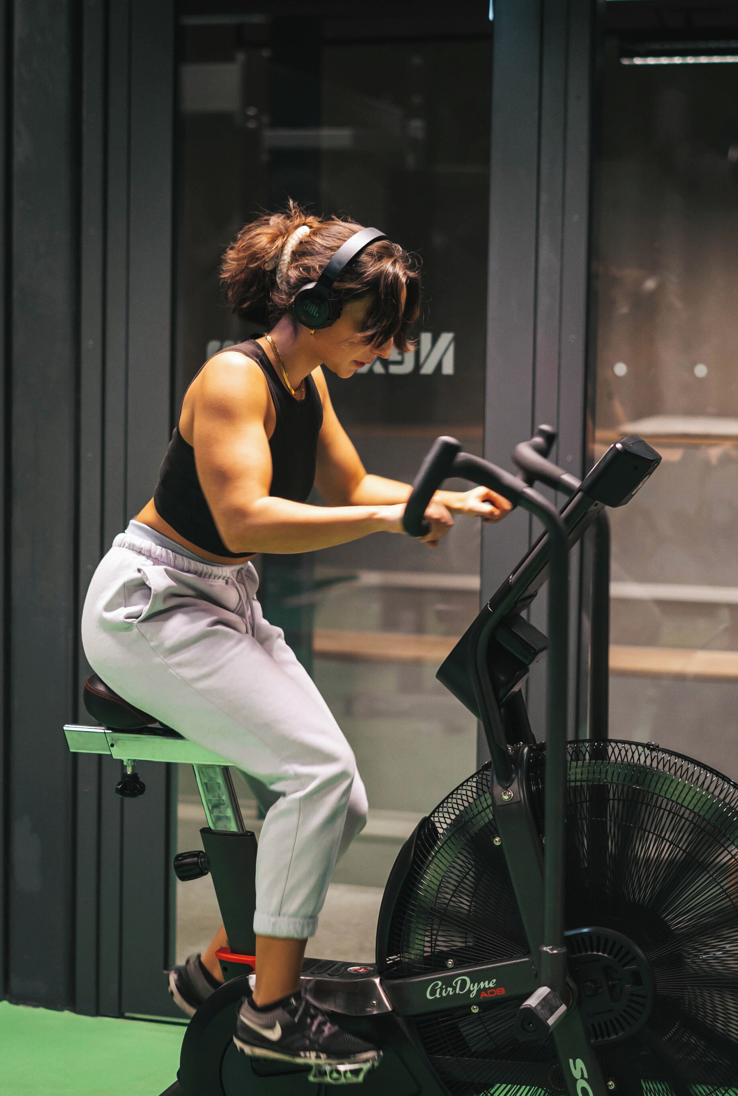 Une personne fait de l’exercice sur un vélo d’appartement dans une salle de sport, avec un grand ventilateur et un mur en verre en arrière-plan.