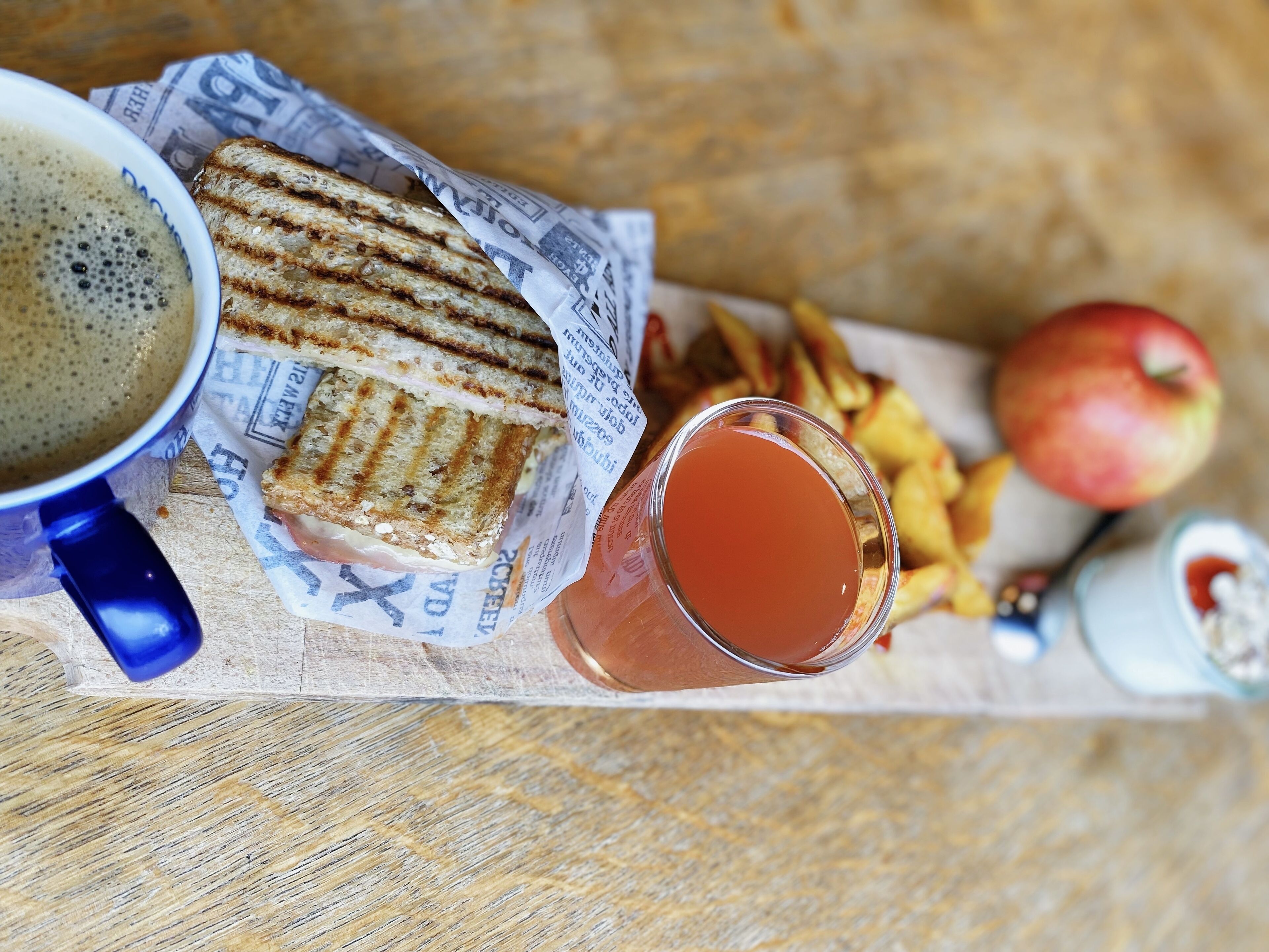 Un sandwich posé sur un journal, un verre de jus de fruits, des frites, une pomme et une tasse de café sur une table en bois.