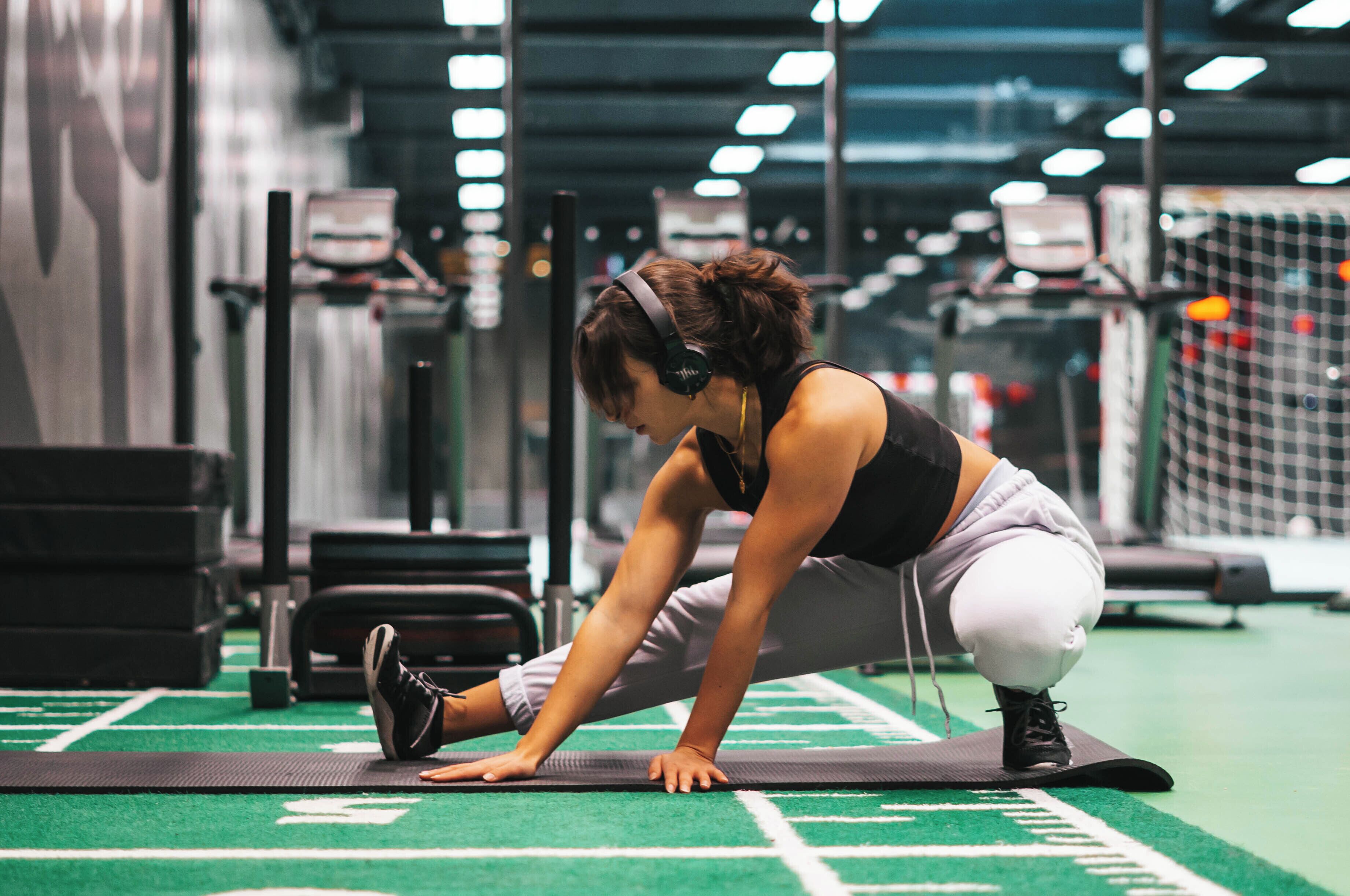 Una persona estirándose sobre una colchoneta en un gimnasio, con equipo de ejercicio al fondo.