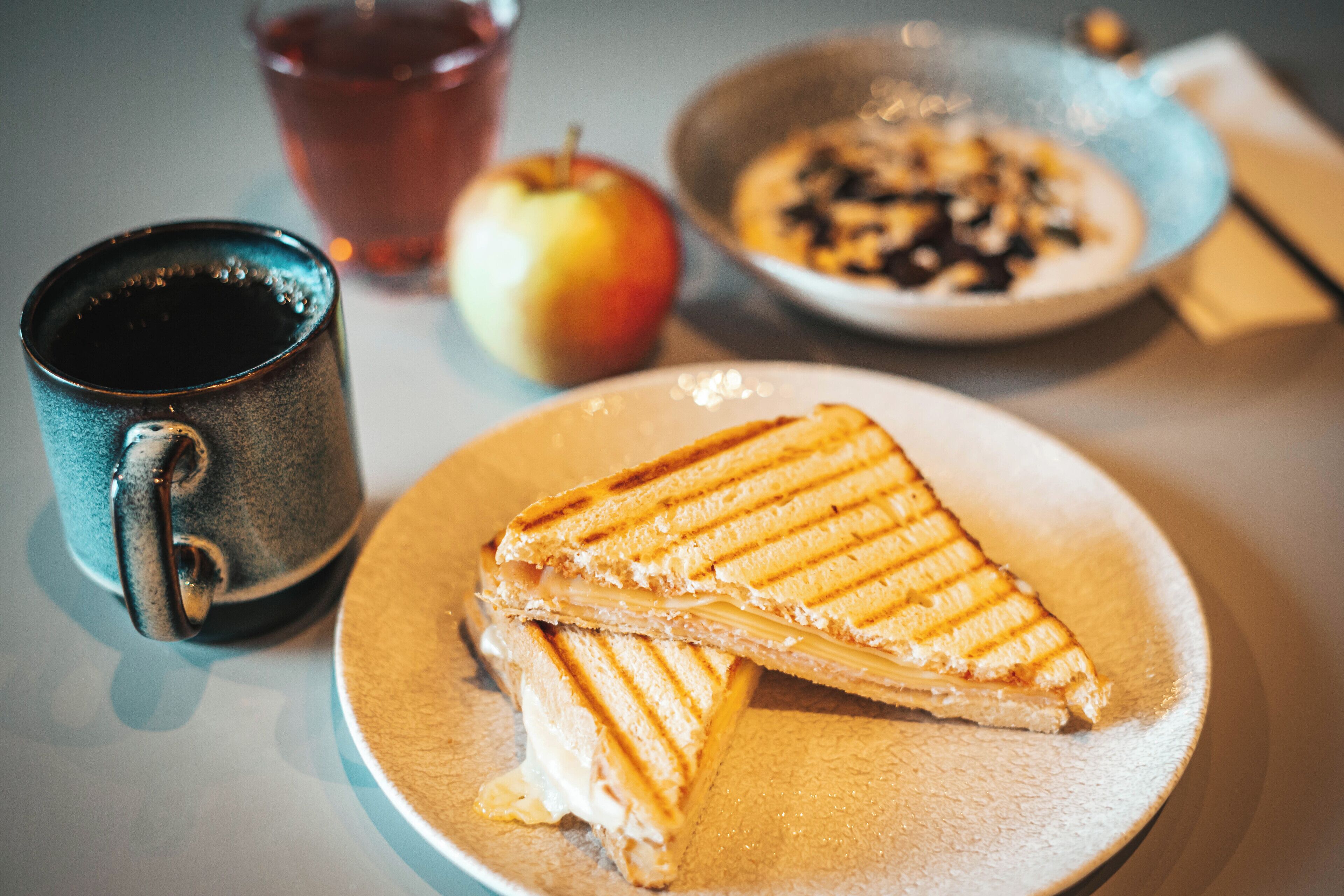 Un plato con un sándwich a la plancha, una taza, una manzana y un tazón de cereal.