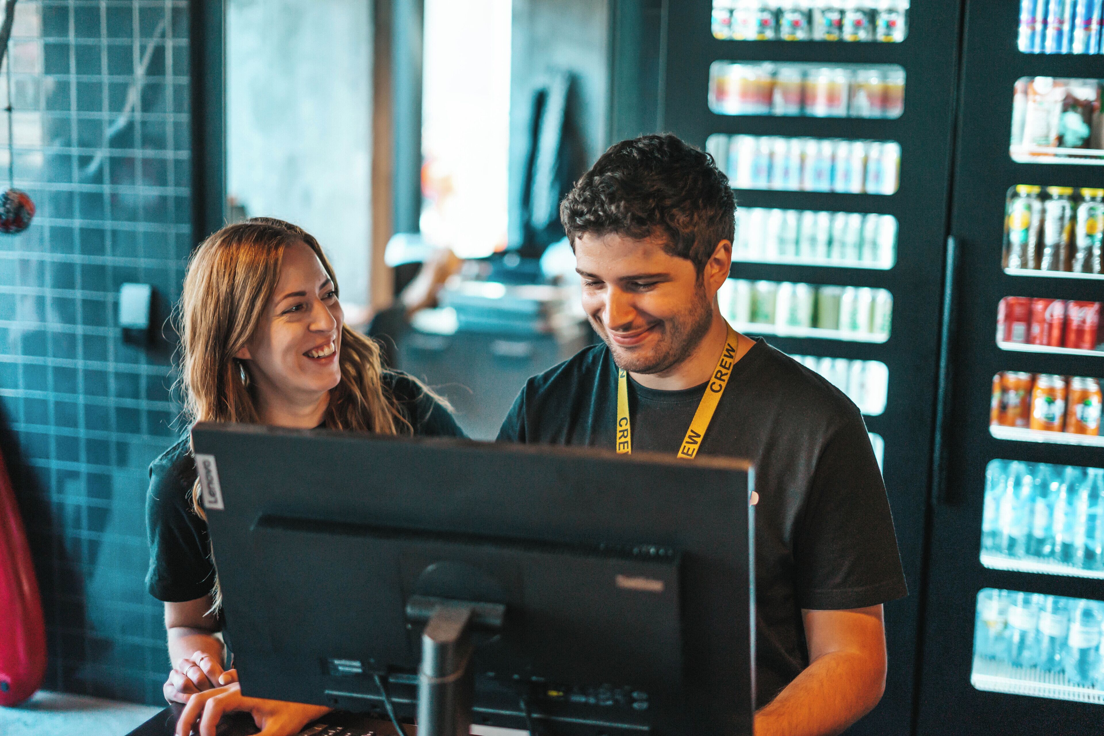 Two people interacting with a computer monitor in a room with vending machines in the background.