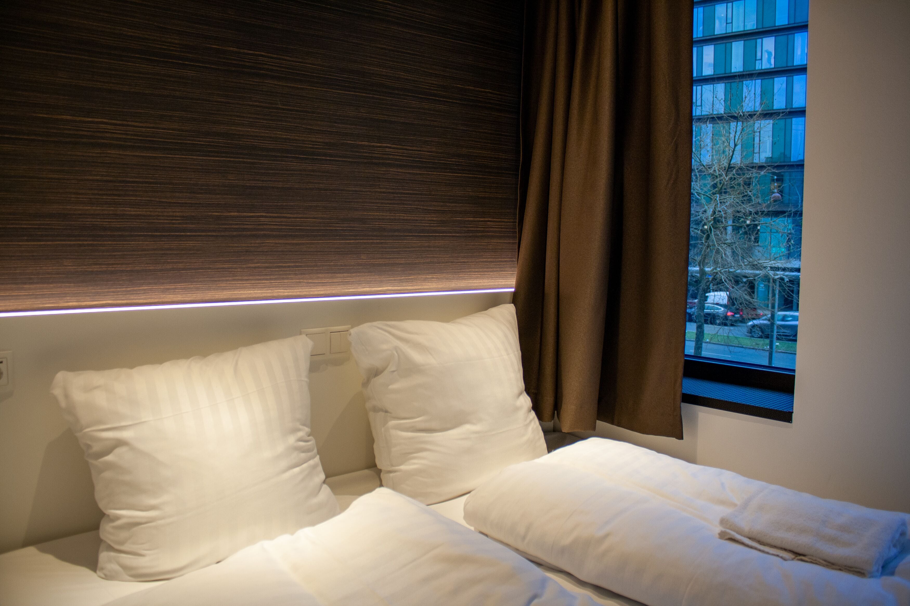 A hotel room with a bed, white pillows, a window with curtains, and a view of the outside.
