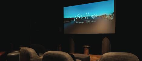 A darkened room with a large screen displaying 'Next House' and 'ARP-Hansen Hotel Group'. There are several upholstered seating areas and a wooden floor.