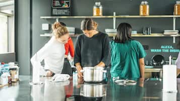 Un área de cocina con tres personas preparando alimentos, una pizarra con los platos del menú y estantes con frascos y platos.