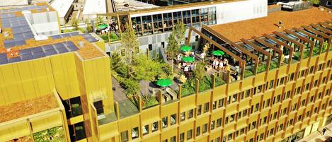 A high-rise building with multiple floors, large windows, and a rooftop garden area with people and umbrellas.