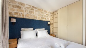 Une chambre d’hôtel avec un mur en pierre, une tête de lit bleue, un lit avec du linge de maison blanc, deux lampes de chevet et une armoire.