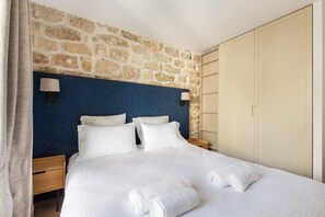A hotel room with a stone wall, a blue headboard, a bed with white linens, two bedside lamps, and a wardrobe. - St Georges - One Bedroom in Paris South Pigalle (Paris)