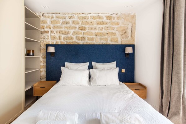 Un lit bien fait, avec du linge de lit blanc, une tête de lit bleue et deux lampes de chevet.
