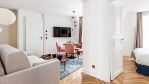Moderni hotellihuone, jossa on sohva, vaaleanpunaisilla tuoleilla varustettu ruokailutila, seinälle asennettu televisio ja puulattia.