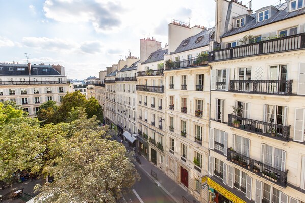 A row of multi-story buildings with balconies, a street in between, and trees on the left.
