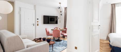 Una moderna camera d'albergo con divano, una zona pranzo con sedie rosa, una TV montata a parete e un pavimento in legno.