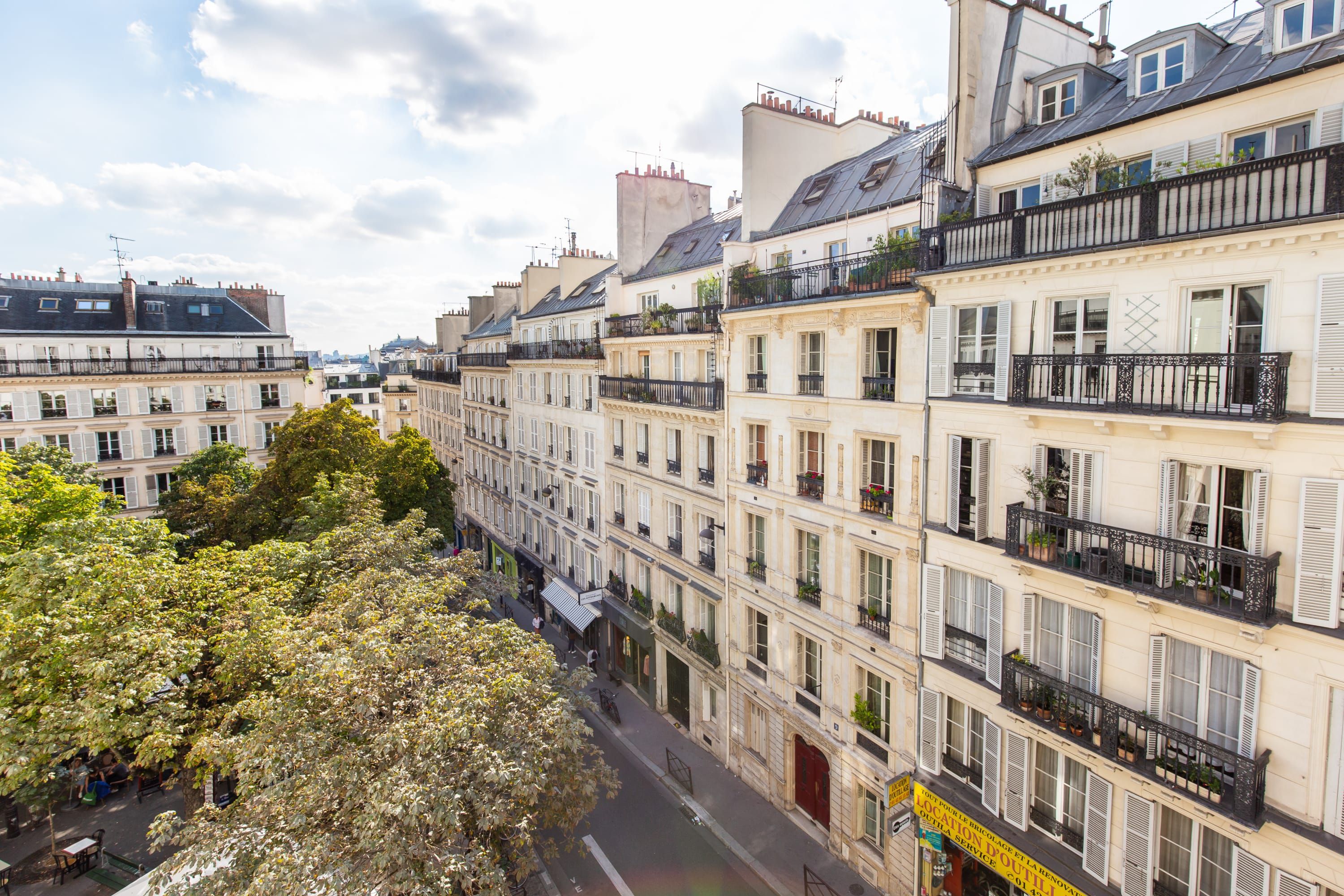 A row of multi-story buildings with balconies, a street in between, and trees on the left.