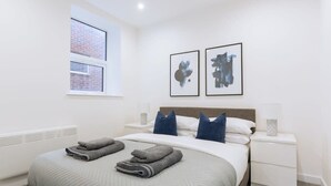 Une chambre moderne avec un grand lit, deux tables de chevet, une fenêtre donnant sur un immeuble en briques et deux œuvres d’art abstraites accrochées au mur.