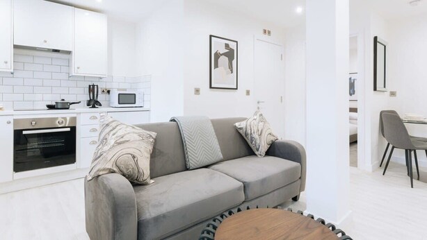 Un espace de vie moderne avec un canapé gris, une table basse en bois, une cuisine avec des meubles blancs et un coin repas avec une chaise grise.