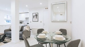 Une cuisine moderne avec des meubles blancs, un coin repas doté d’une table en verre et un grand miroir au mur.