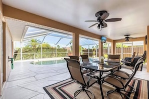Area pranzo a bordo piscina con tavolo e sedie, ventilatore a soffitto e vista sulla piscina e sul verde.