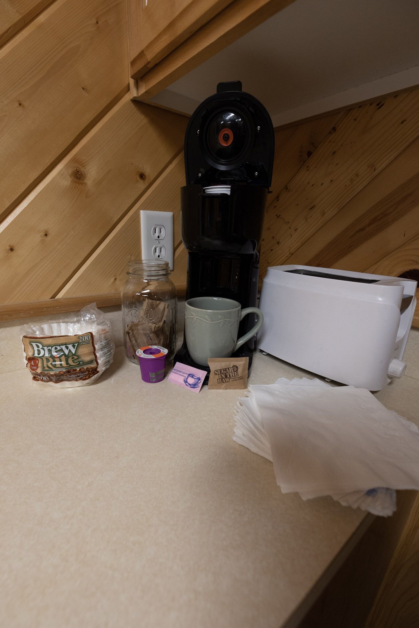 A kitchen counter with a coffee maker, a toaster, a mug, a jar, a bag of coffee, and a packet of tea.