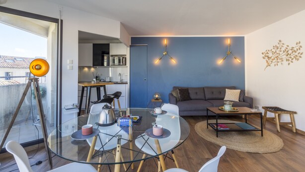 A modern living room with a glass dining table, a sofa, and a coffee table. There is a kitchen in the background with a microwave and a kettle. - Terrasse en ville - Première conciergerie (Montpellier)