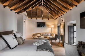 A modern bedroom with a wooden ceiling, a large bed with white bedding, a sofa, a dining table, and a TV mounted on the wall.