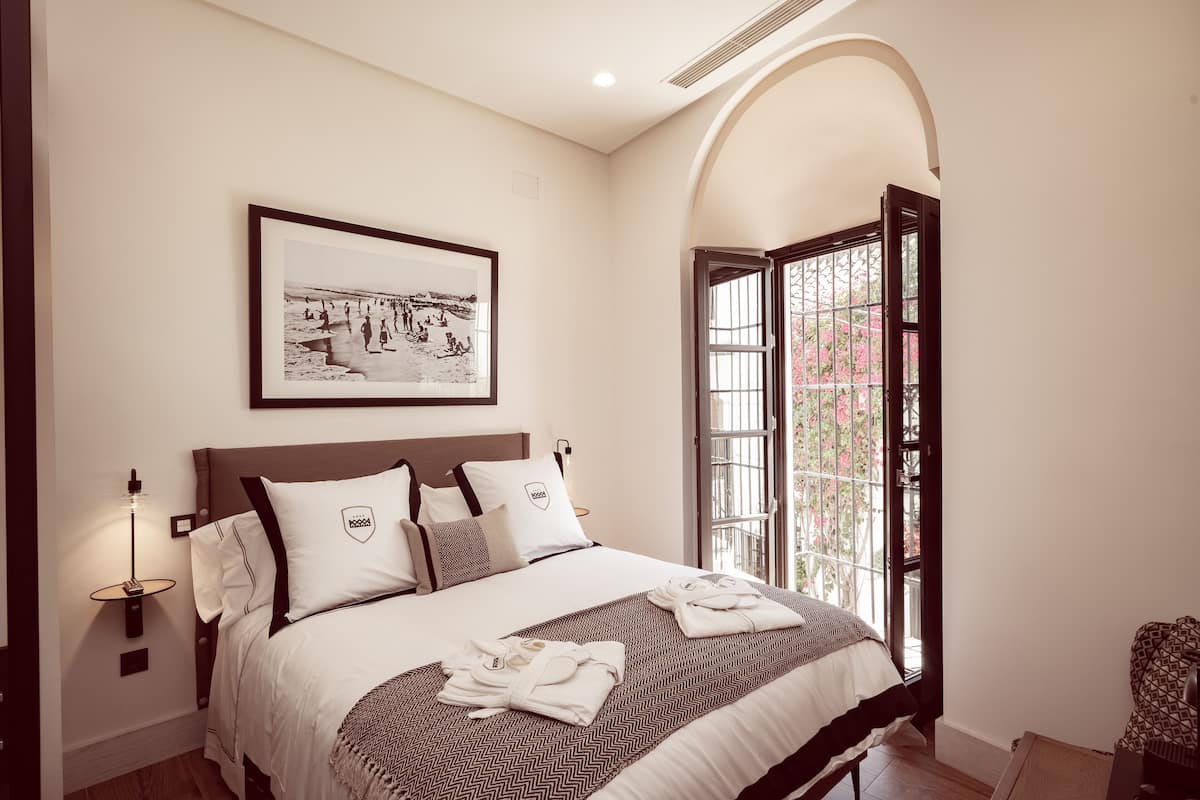 Una camera d'albergo con un letto grande, un quadro appeso al muro, una finestra con vista su fiori rosa e un comodino con una lampada.