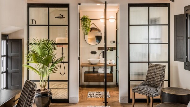 A modern bedroom with a sloped ceiling, a large bed, a sitting area with a chair, a potted plant, and a bathroom with a mirror and sink. - Hotel Maison Ardois La Ciudadela Marbella (Marbella)