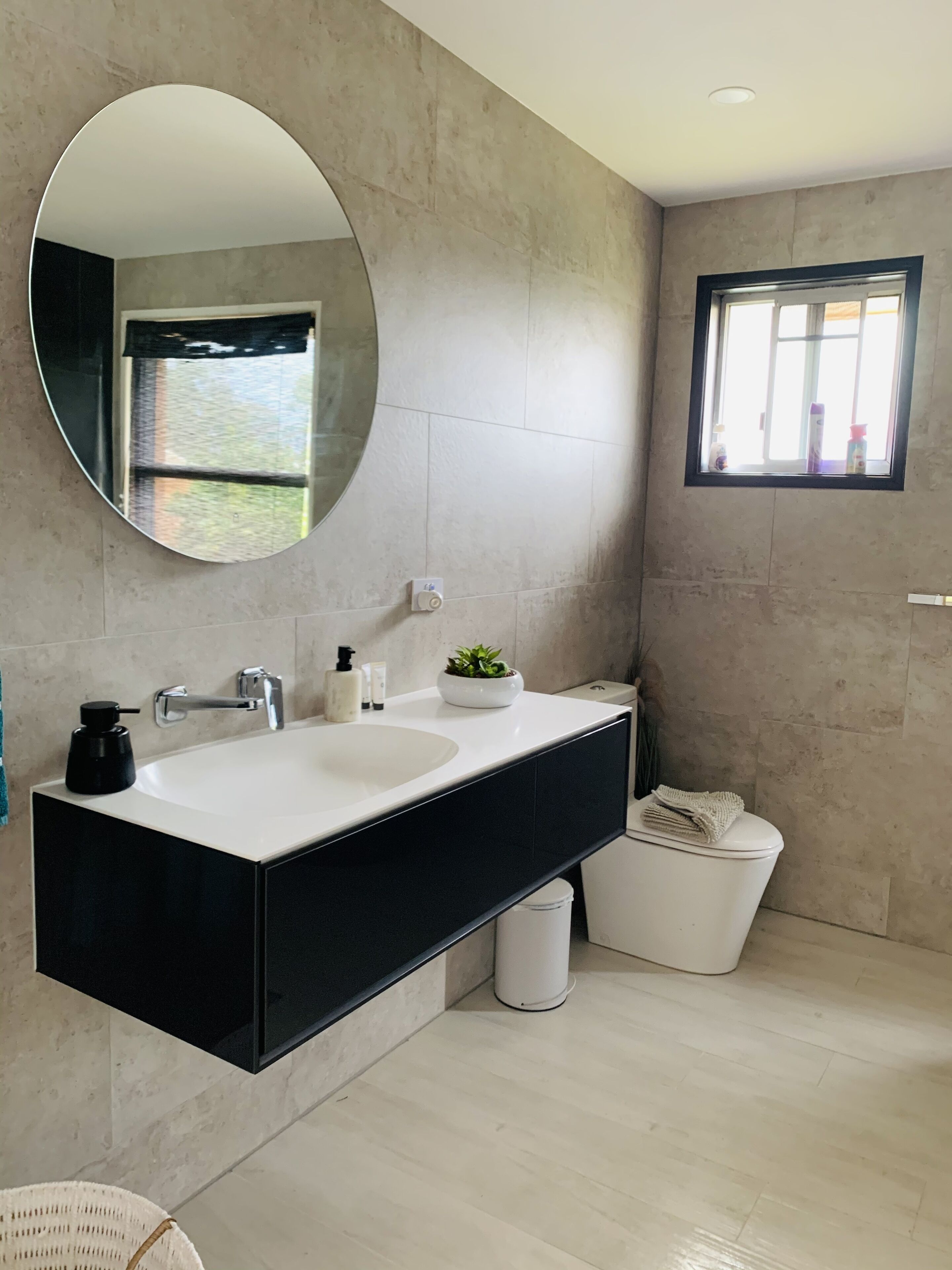 A modern bathroom with a double sink vanity, a round mirror, a small window, and a trash bin.