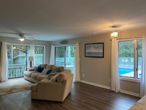 Un salon spacieux avec un canapé beige, un ventilateur de plafond et de grandes fenêtres donnant sur une piscine et une terrasse.