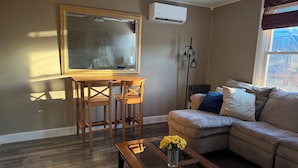 A living room with a wooden table, a sofa, a glass coffee table with flowers, a painting on the wall, and an air conditioning unit. - The Steel City Loft  (Bethlehem)