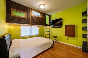 A room with a bed, a TV mounted on the wall, a window with blinds, and a brick wall design.