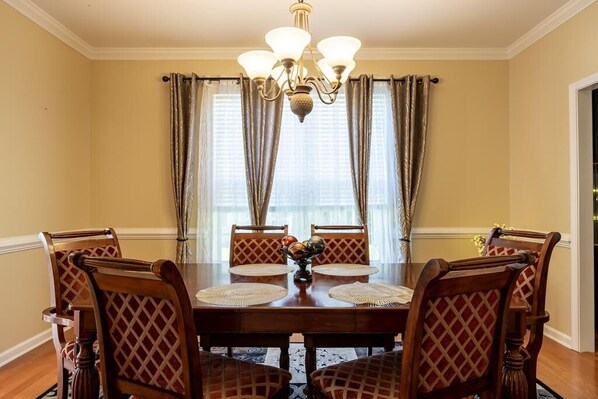 A dining room with a wooden table, six chairs, a chandelier, and a window with curtains.