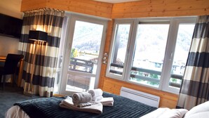 A hotel room with a bed, a desk, and a view of a mountainous landscape through large windows.