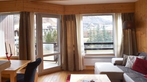 A room with a wooden interior, a sofa, a table, and a view of mountains through large windows.