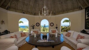 A spacious living room with a round coffee table, white sofas, and a large arched window offering a view of the ocean.