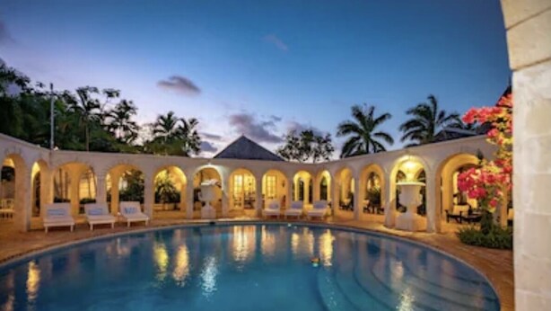 A swimming pool with lounge chairs and arched structures, surrounded by palm trees and flowers.