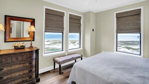 Een slaapkamer met een bed, een dressoir, een bankje en grote ramen met uitzicht op het strand.
