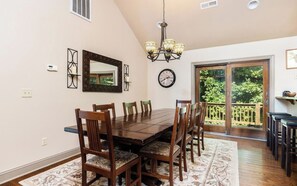A dining room with a wooden table and chairs, a chandelier, a clock, and a view of greenery through a glass door.