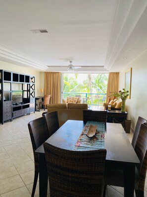 A spacious living room with a large dining table, comfortable seating, a TV, and a balcony with a view of palm trees.