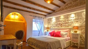 Un dormitorio con pared de piedra, vigas de madera, cama con ropa de cama rosa y blanca, una mesita de noche con lámpara y una silla.