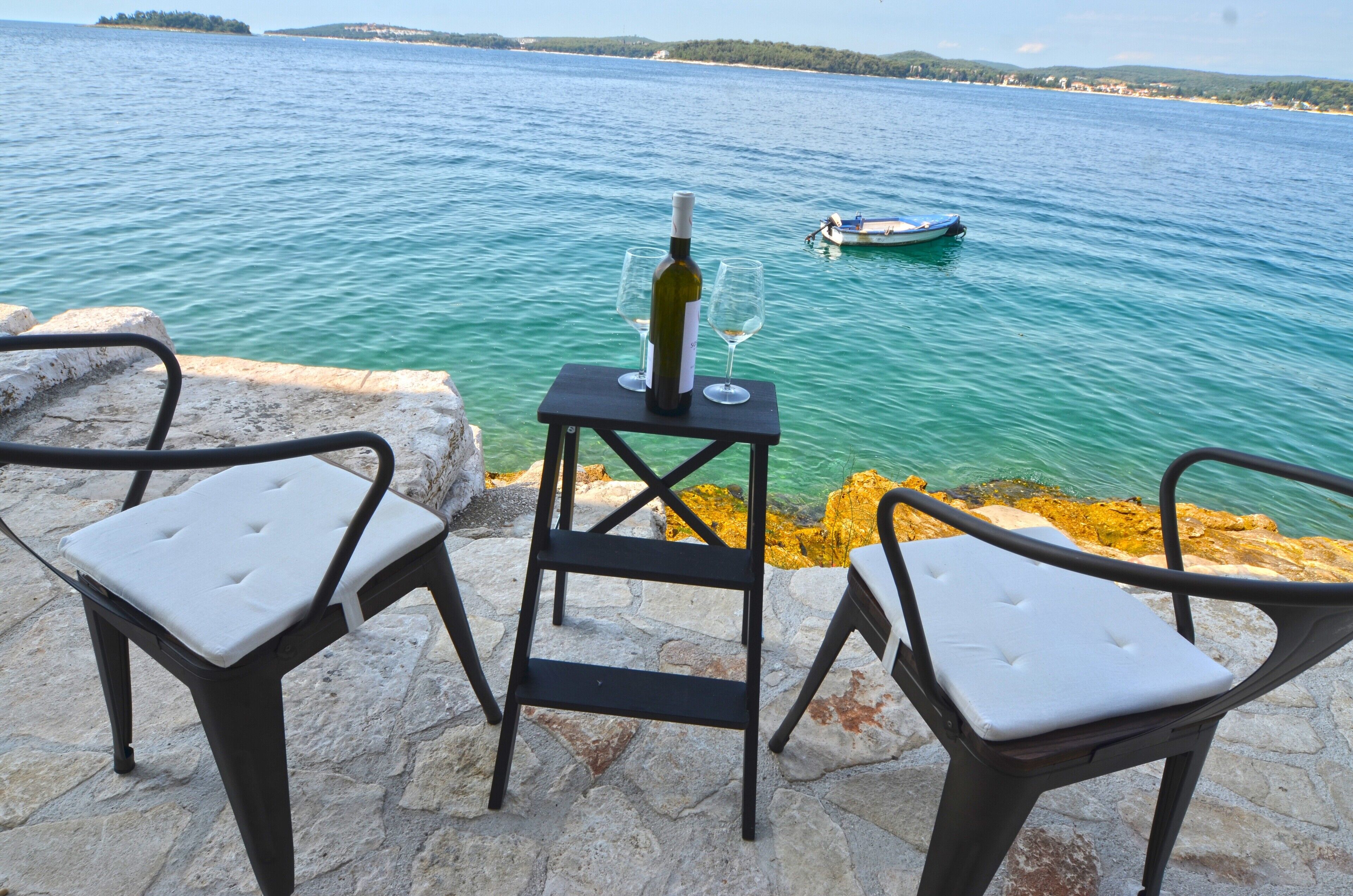 Une table avec une bouteille et des verres, donnant sur une étendue d’eau où se trouve un bateau.
