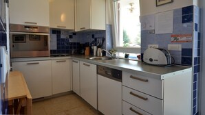 A modern kitchen with white cabinets, a built-in oven, a dishwasher, and a window with a view of greenery outside. - Alma Borik Apartment with balcony (Rovinj)