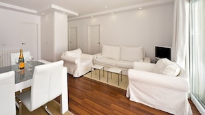 A modern, white-themed living room with a sofa set, a dining table with chairs, and a TV.