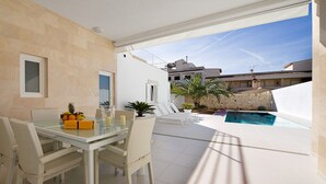 A modern outdoor dining area with a glass table and wicker chairs, a pool, and a view of the surrounding buildings.