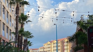 A hotel corridor with palm trees, string lights, and a clear sky.