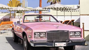 A pink Cadillac convertible parked in a sunny outdoor area with palm trees and a white building with a circular structure in the background.