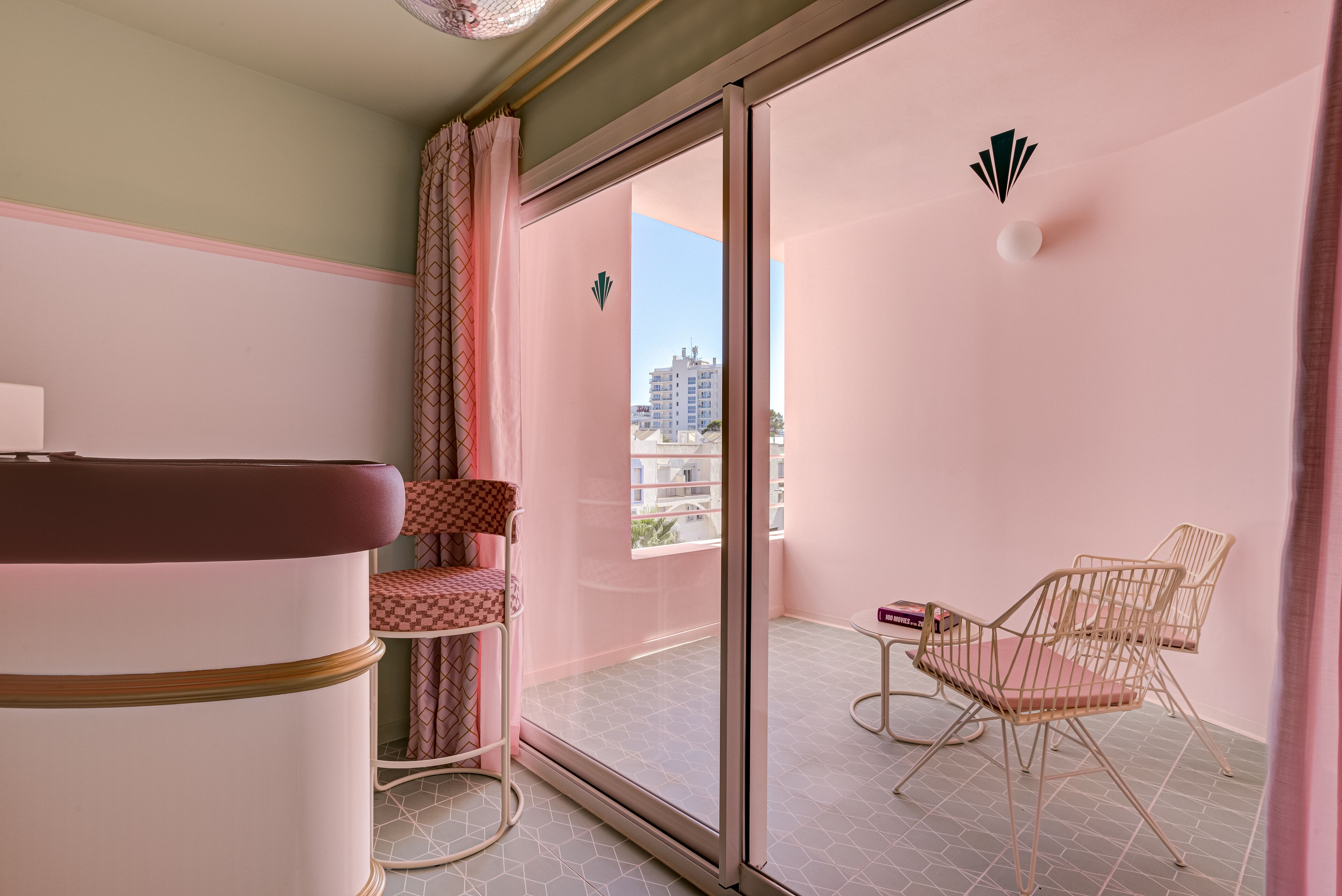 A balcony with a pink interior, a cushioned chair, a small table, and a view of buildings.
