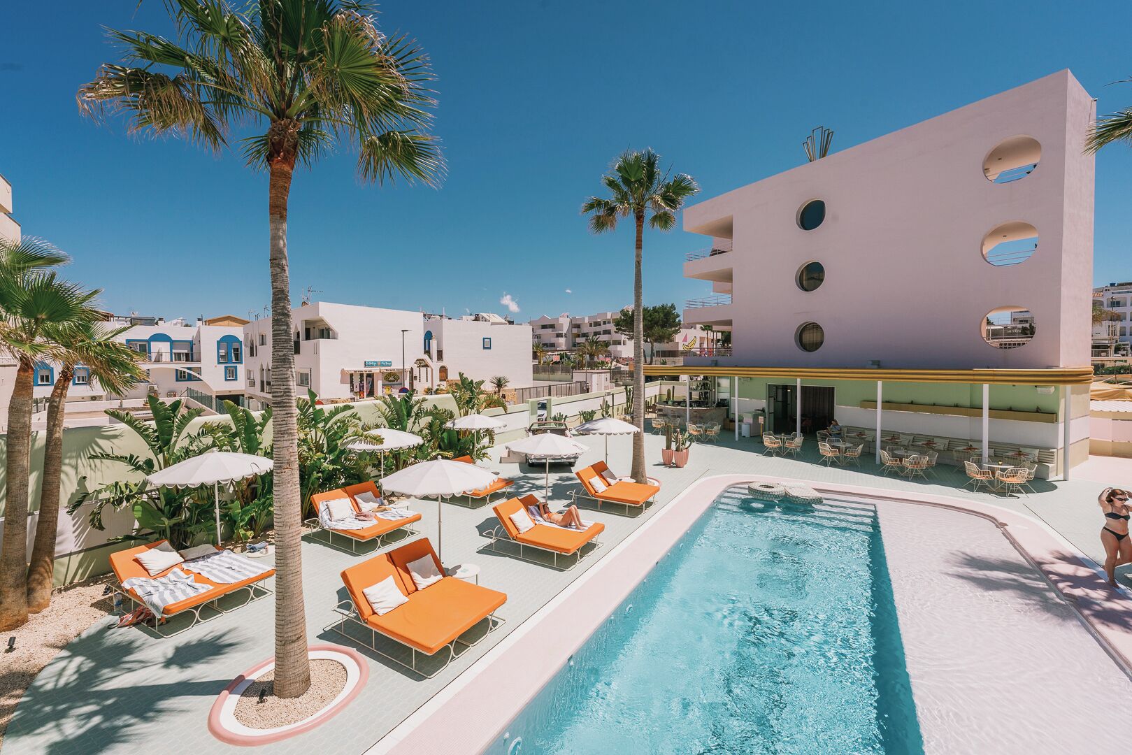 Zona de piscina con sillones de descanso y sombrillas, rodeada de palmeras y un edificio con ventanas circulares.