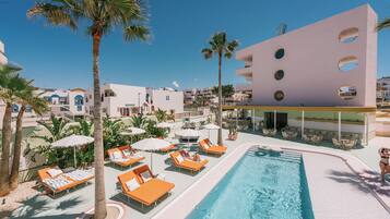 Zona de piscina con sillones de descanso y sombrillas, rodeada de palmeras y un edificio con ventanas circulares.